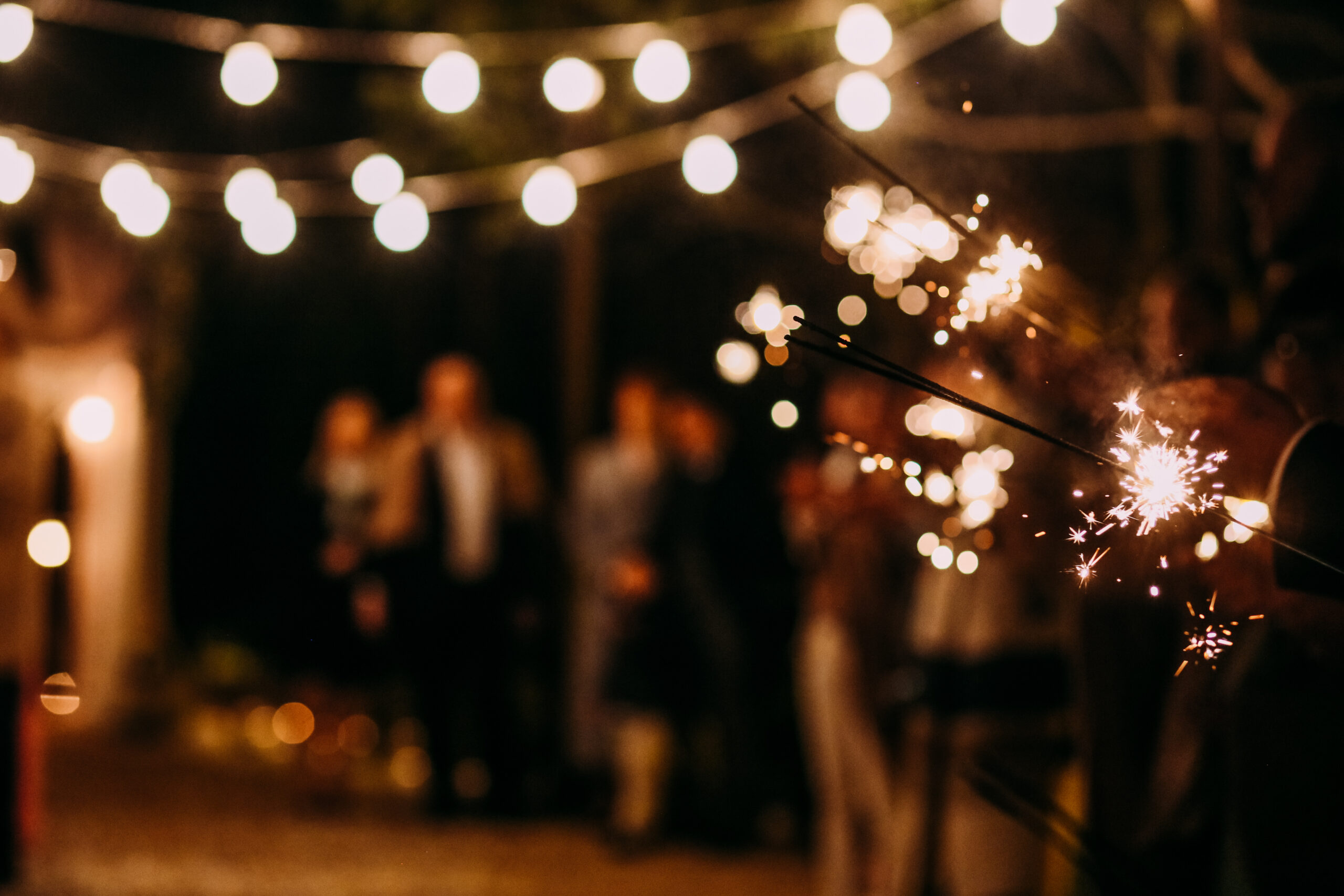 Nighttime celebration with warm string lights and a hand holding a lit sparkler in the foreground; blurred guests in the background.